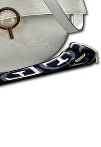 Witte leren schoudertas met een ronde vorm, voorzien van een gouden hardware ring en een gestreepte marineblauwe schouderband met witte accenten.