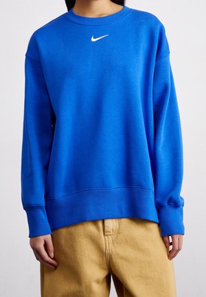 Personne portant un sweat Nike bleu vif avec un logo blanc et un pantalon beige ample, debout devant un fond uni.