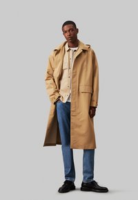 Beige lang frakk med en sammenleggbar krage, brystlomme og store frontlommer, båret over en lys skjorte og blå jeans. Svarte sko.