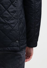 Svart vadderad jacka med diamantmönster, metallknappar och en slät textur, visad bakifrån med grå jeans.