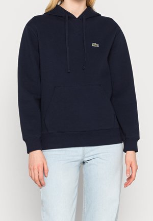 Person iført en marineblå hættetrøje med en frontlomme og lille grønt krokodillelogo, kombineret med lyseblå jeans, stående med armene afslappede.