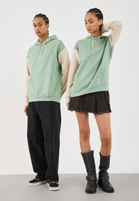 Due modelli indossano felpe verdi e beige; uno abbinato a pantaloni larghi neri e sneakers, l'altro con una gonna nera e stivali neri.
