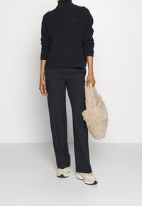 Persona che indossa un maglione a collo alto a coste nero e pantaloni neri, con una borsa tote in pelliccia beige, indossando sneakers beige chiaro.