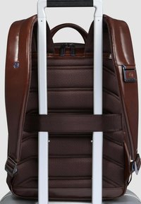 Zaino in pelle marrone agganciato a una maniglia argentata di un trolley; presenta una texture imbottita, doppia chiusura lampo e dettagli con logo. Finitura liscia.