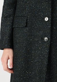 Manteau en tweed noir avec une finition texturée, comportant une poche, quatre boutons noirs et de subtiles éclats colorés répartis dans le tissu.
