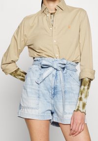 Světle béžová košile na knoflíky s logem, v kombinaci s vysokými světle modrými džínovými šortkami s páskem a kapsami.