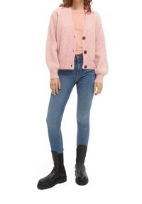 Cardigan de canalé rosa claro con mangas largas, cierre de botones y botones negros. Combinado con una camisa rosa y jeans ajustados azules, y botas negras.