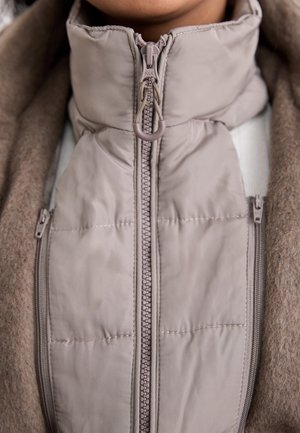Beige wattierte Jacke mit Reißverschluss und hohem Kragen, getragen unter einem braunen Wollmantel an einer Person, die den vorderen Mittelbereich und den Hals zeigt.