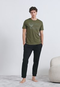 Olivzaļš kokvilnas t-krekls ar "EMPORIO ARMANI" logotipu, kopā ar melniem šauriem biksēm no mīkstas auduma. Modelis stāv basām kājām uz paklāja.