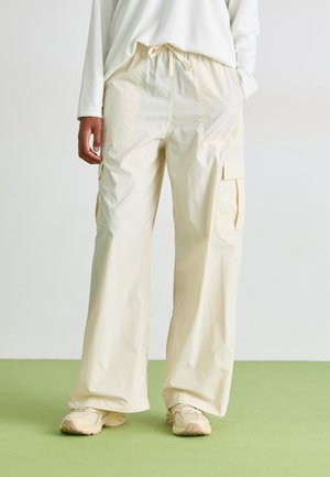 Pantaloni cargo beige chiaro con vestibilità ampia, vita elasticizzata, due tasche laterali e tessuto testurizzato. Indossati con scarpe da ginnastica beige su un pavimento verde.
