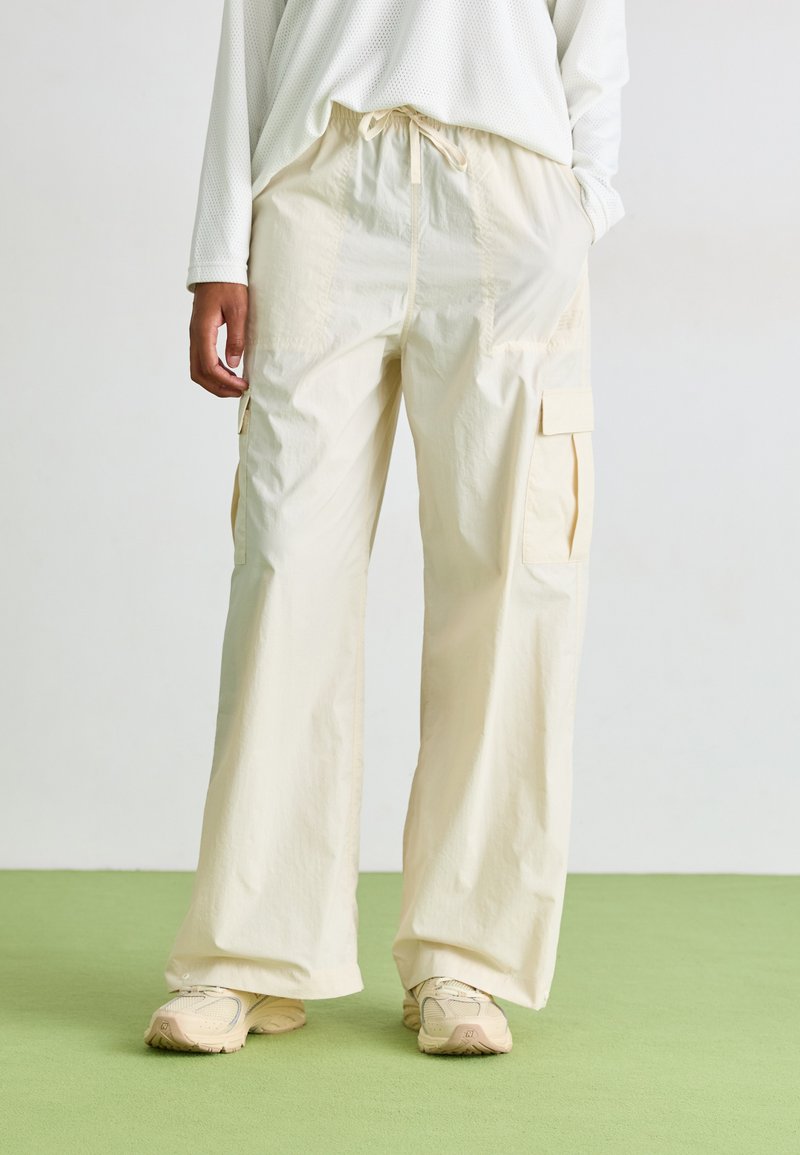 Light beige cargo pants with a loose fit, elastic waistband, two side pockets, and textured fabric. Worn with beige sneakers on green floor.
