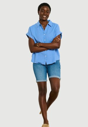 Kobieta stoi ze skrzyżowanymi rękami, ubrana w jasnoniebieską, krótką koszulę na guziki, dżinsowe szorty i beżowe buty wsuwane.