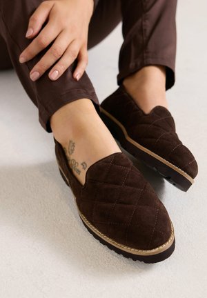 Personne portant des chaussures marron foncé matelassées sans lacets avec des semelles beiges et un pantalon marron, montrant un petit tatouage à la cheville sur une peau claire.