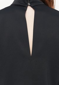 Garment in tessuto nero con colletto alto e apertura a forma di chiave sul retro, fissata da un bottone al collo. Texture liscia.