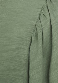 Grønt strikket stof med en blød tekstur. Har sammensamlede detaljer ved skulderen, der skaber subtil volumen og dybde.