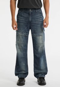 Blauwe denim cargobroek met een losse pasvorm, meerdere voor- en zijzakken, een vervaagde textuur en een knoopsluiting. Draag met zwarte schoenen.