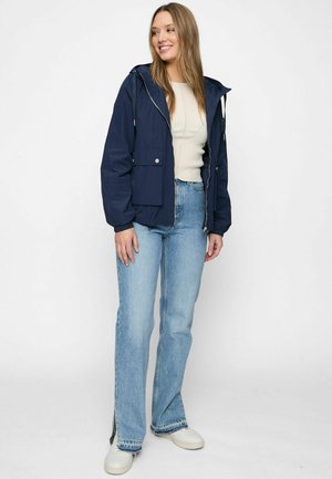 Jeune femme portant une veste à capuche bleu marine, un pull beige, un jean bleu clair avec des fentes latérales, et des baskets blanches, debout et souriante.