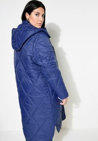 Chaqueta larga acolchada en color navy con capucha, que presenta un patrón de diamantes y una textura brillante. Cierre con cremallera en el frente y bolsillos laterales visibles.