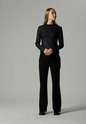 Jonge vrouw in een zwarte textuur lange mouw top en zwarte broek staat met haar handen voor zich samengevouwen en kijkt naar rechts tegen een effen grijze achtergrond.