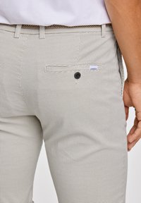 Pantalones texturizados gris claro que presentan un pequeño bolsillo trasero, un cierre de botón y un cinturón trenzado, mostrando un patrón sutil.