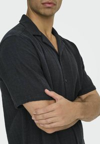 Schwarzes kurzärmeliges Hemd aus strukturiertem Stoff mit einem Reverskragen, ausgestattet mit einem einzelnen Knopf und einer lockeren Passform.