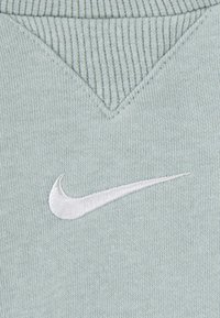 Šviesiai žalias medvilnės audinys su šonkaulinės tekstūros, turintis V formos iškirpimą ir baltą siuvinėtą Nike logotipą apatinėje kairėje pusėje.