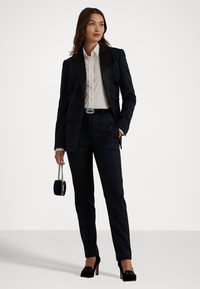 Traje de cuadros negros con chaqueta de doble botón ajustada, camisa blanca, cinturón negro y tacones de aguja. Incluye un pequeño bolso de mano azul.