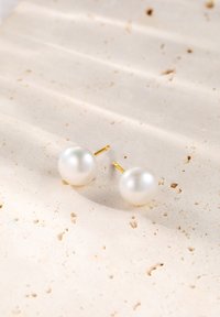 Orecchini a bottone con perle dal design sferico e liscio in bianco, dotati di perni in tonalità oro. Posizionati su una superficie testurizzata di beige chiaro.