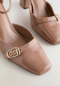 Beige kiilakorkoiset kengät joka on valmistettu kiiltävästä nahasta, neliömäisellä kärjellä ja koristeellisella kultaisella soljella, joka kulkee päällä.