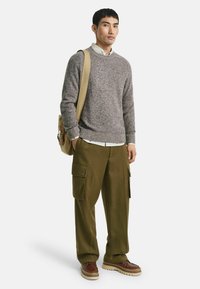 Grauer Strickpullover, beige Hemd mit Kragen darunter, olivgrüne Cargo-Hosen mit Taschen, braune Schuhe und eine khakifarbene Umhängetasche.
