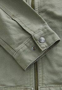 Close-up of an olive green jacket sleeve with visible stitching, a metal snap button, and a partially visible brass zipper.