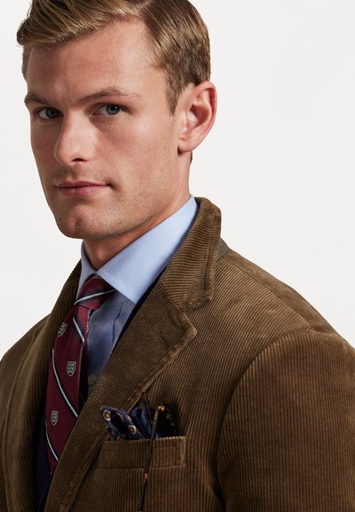 Blazer en velours côtelé marron avec revers crantés, porté sur une chemise habillée bleu clair, associé à une cravate bordeaux à motifs subtils et un mouchoir de poche.