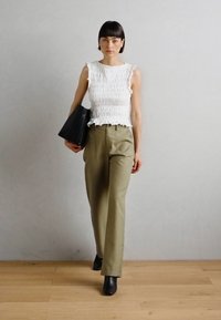 Haut sans manches blanc texturé avec des volants, associé à un pantalon taille haute vert olive et des bottines noires, tenant un sac noir.