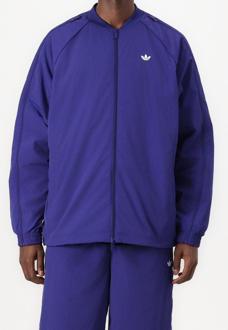 Lila zip-up jacka i lättviktig tyg, med elastiska muddarm, ståkrage och en liten vit logotyp på bröstet.