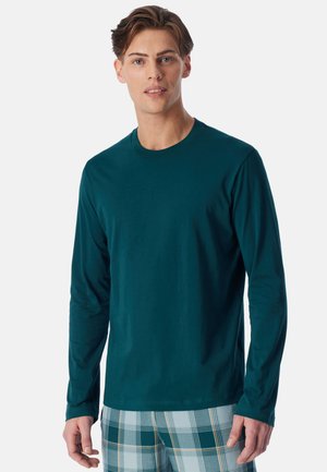 Jonge man met een donkergroen overhemd met lange mouwen en blauwgroene geruite broek, staand tegen een effen witte achtergrond.