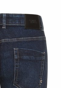 Jean en denim bleu foncé avec une apparence délavée, arborant une étiquette de marque en cuir noir et une poche arrière avec des coutures renforcées.