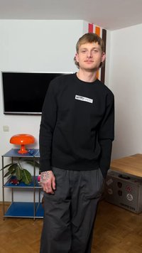 Nuori mies, jolla on tatuointeja kädessä, pukeutunut mustaan collegepaitaan ja harmaisiin housuihin, seisomassa huoneessa, jossa on oranssi lamppu, kasvi ja seinälle kiinnitetty televisio.