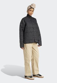 Femme portant un turban beige, une veste matelassée noire, un pantalon beige et des baskets noir et blanc, se tenant devant un fond uni.