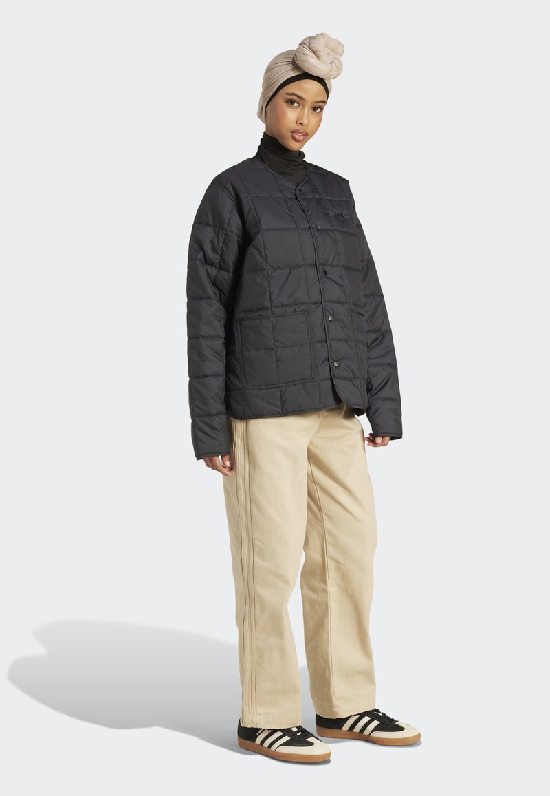 Femme portant un turban beige, une veste matelassée noire, un pantalon beige et des baskets noir et blanc, se tenant devant un fond uni.