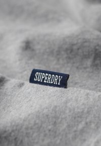 Grijs stof met een gestructureerd oppervlak. Bevat een marineblauwe label met "SUPERDRY" in witte geborduurde letters. Eenvoudig en strak ontwerp.