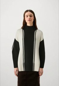 Sieviete stāv, vērsta uz priekšu, ģērbta melnbaltā augsta apkakle džemperī ar vertikālām adījuma svītrām un garām piedurknēm.