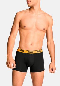 Schwarze Boxershorts mit einem gelben Puma-Bund, in enganliegendem Design und mittellanger Länge bis zum Oberschenkel. Glatte Textur und Baumwollmaterial.