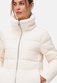 Doudoune en crème avec un col haut, un design matelassé horizontal et une texture douce ; elle dispose d'une fermeture éclair sur le devant et de poches latérales.