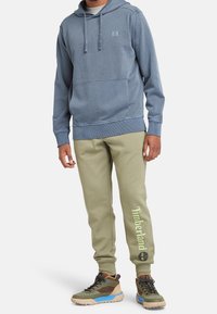 Sudadera con capucha de algodón azul con bolsillo frontal, combinada con pantalones de chándal oliva que tienen el logotipo de "Timberland" en verde claro, y zapatillas en tonos beige y azul.