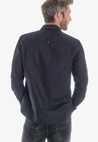 Camisa de manga larga azul marino en mezcla de algodón con cuello abotonado. Presenta un pequeño logotipo rojo y blanco en la parte posterior. Textura suave.