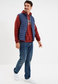 Chaleco acolchado navy sobre una sudadera roja con capucha, vaqueros azules y zapatillas blancas. El chaleco presenta un acolchado horizontal y un cierre de cremallera selectivo.