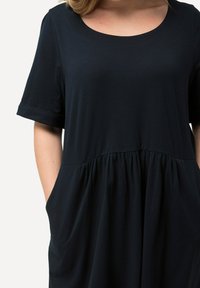 Tummansininen mekko, jossa on lyhyet käännetyt hihat, pyöreä pääntie, vyötärösauma ja kellotettu hame, jossa on sivutaskut. Pehmeä ja joustava kangas.