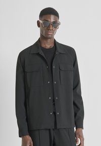 Zwarte jas met twee voorzakken, knoopsluiting en een puntige kraag. Draagt over een zwart shirt, gecombineerd met grijze zonnebril.