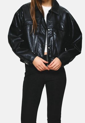 Kvinde iført en sort cropped faux læderjakke over en hvid top, lukker lynlåsen, kombineret med sorte bukser.