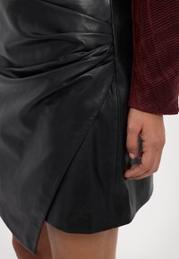 Gonna mini in pelle nera con design arricciato e orlo diagonale. Tessuto liscio con una leggera lucentezza. Mano appoggiata sul bordo della gonna.